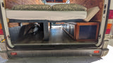 Cozy rv rental camper van interior: raised bed with pillows, under-bed storage cabinets, wood-paneled walls, and laminate flooring.