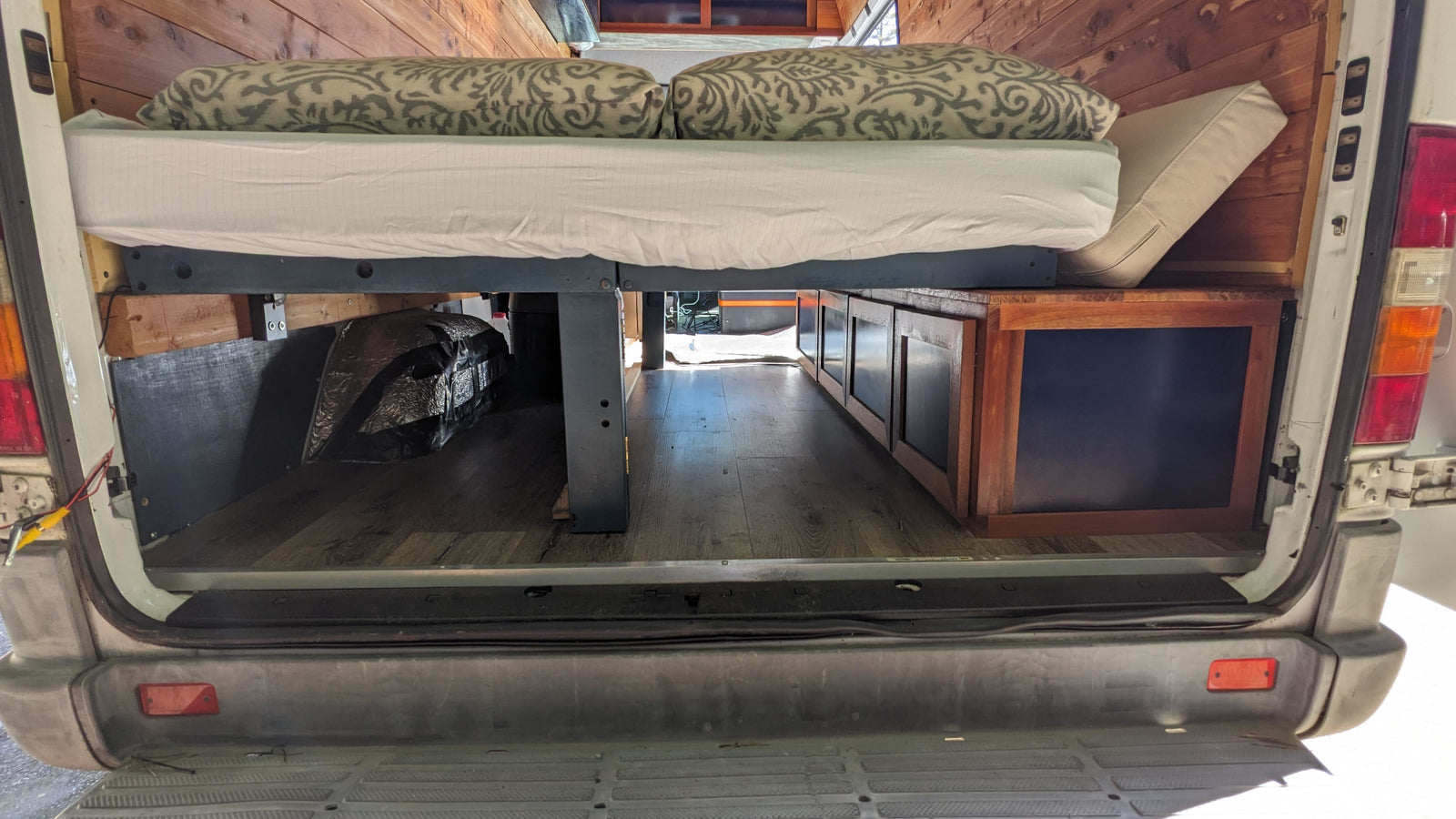 Cozy rv rental camper van interior: raised bed with pillows, under-bed storage cabinets, wood-paneled walls, and laminate flooring.