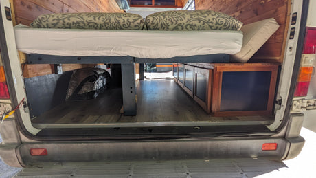 Cozy rv rental camper van interior: raised bed with pillows, under-bed storage cabinets, wood-paneled walls, and laminate flooring.