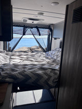 Experience an rv rental! View from the bed shows a serene lake, rocks, and RV interior details like AC, netting & floor.