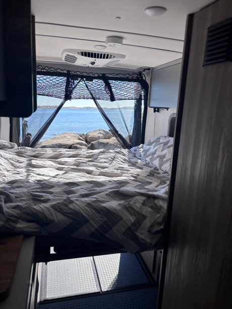 Experience an rv rental! View from the bed shows a serene lake, rocks, and RV interior details like AC, netting & floor.