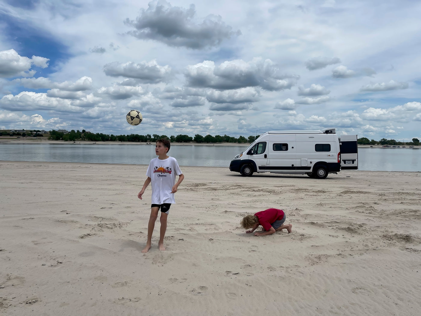 Kids play on a sandy beach by a lake with a white RV rental. One boy kicks a soccer ball, another digs in sand under cloudy skies. — photo 13