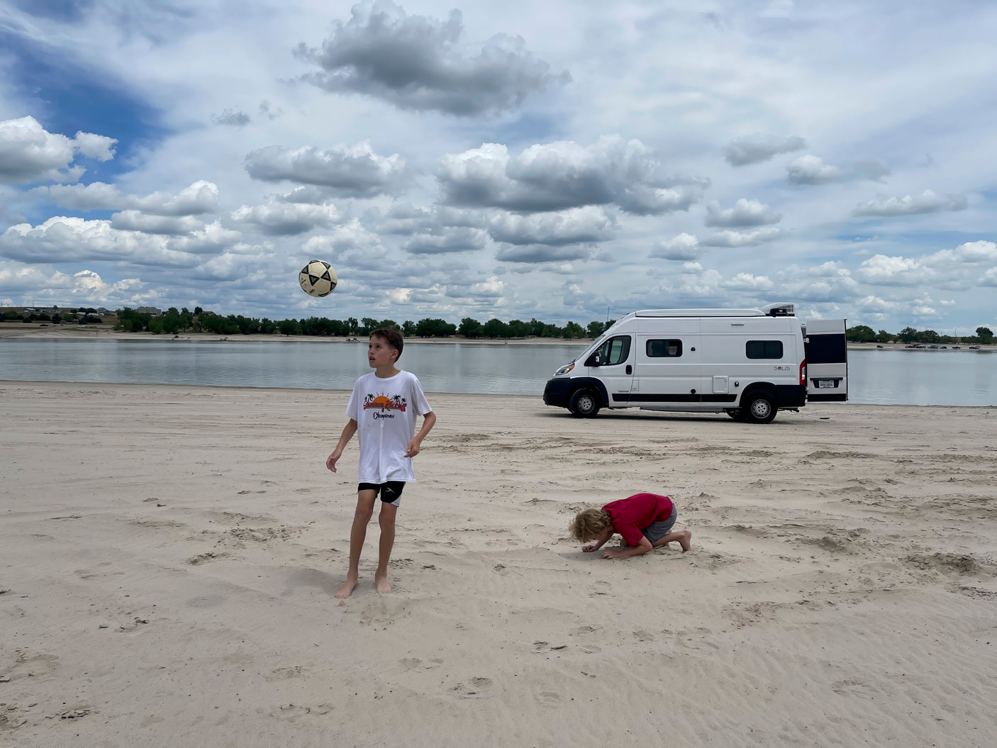 Kids play on a sandy beach by a lake with a white RV rental. One boy kicks a soccer ball, another digs in sand under cloudy skies.