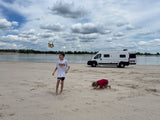Kids play on a sandy beach by a lake with a white RV rental. One boy kicks a soccer ball, another digs in sand under cloudy skies.