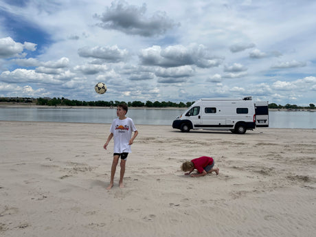 Kids play on a sandy beach by a lake with a white RV rental. One boy kicks a soccer ball, another digs in sand under cloudy skies.