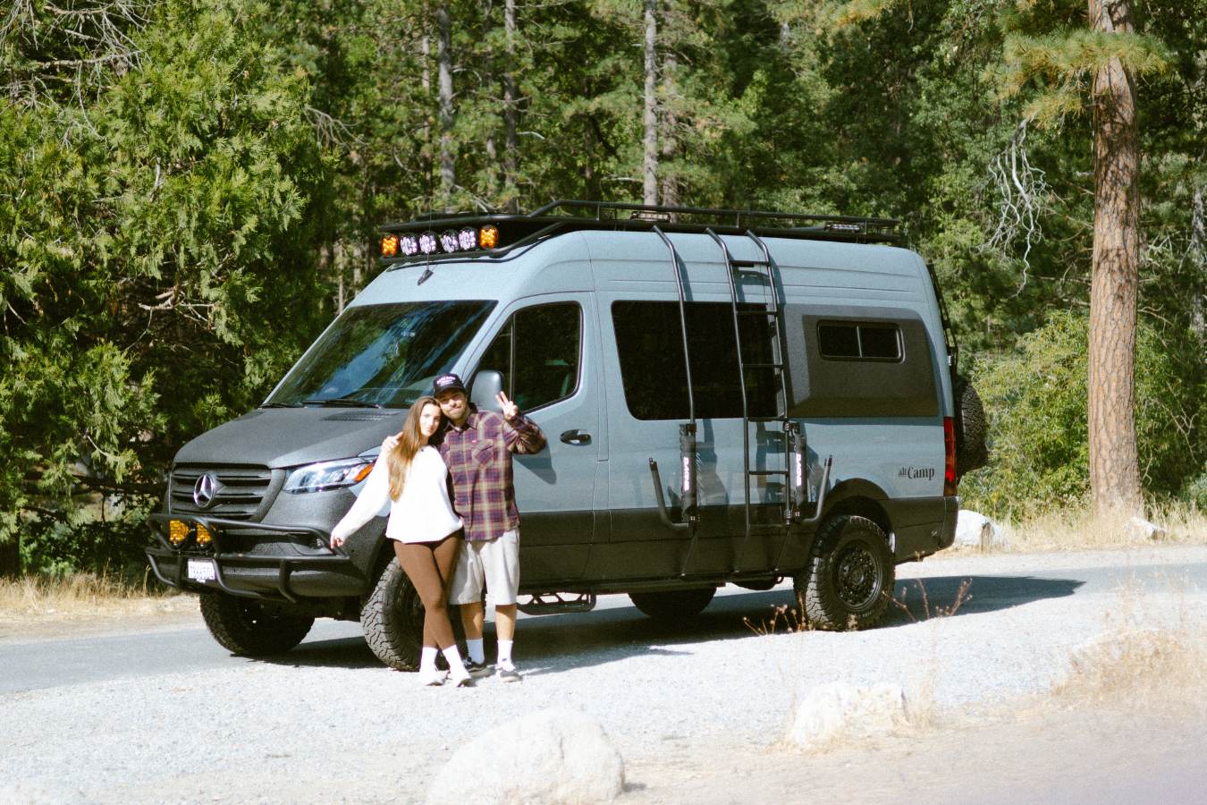 A smiling couple next to their rugged gray RV rental camper van, parked in a scenic wooded outdoor setting for adventure.