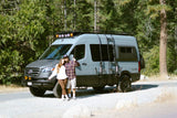 A smiling couple next to their rugged gray RV rental camper van, parked in a scenic wooded outdoor setting for adventure.