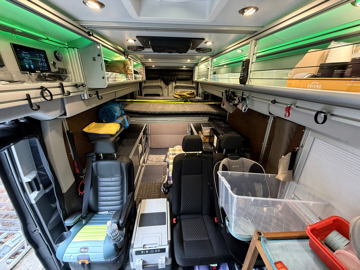 This RV rental interior features a functional van conversion with front seating, a child booster, a bed, and extensive storage with green LED lighting.