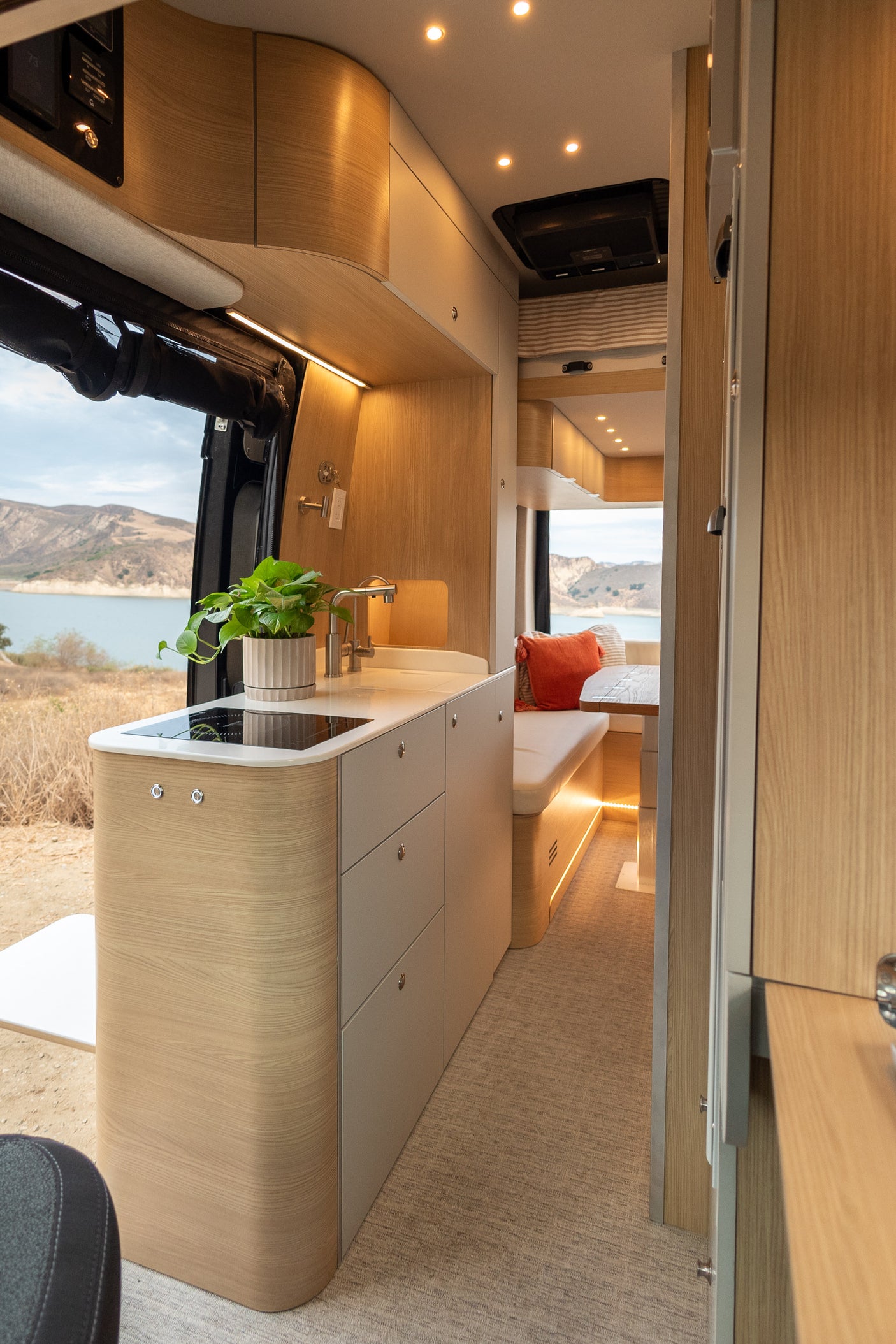 Inside a bright RV rental: a modern kitchenette with a plant, next to a cozy dining area, all overlooking a serene lake.