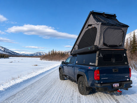 Blue overlanding pickup truck with pop-up camper (RV rental) on a snowy winter road, mountains ahead.