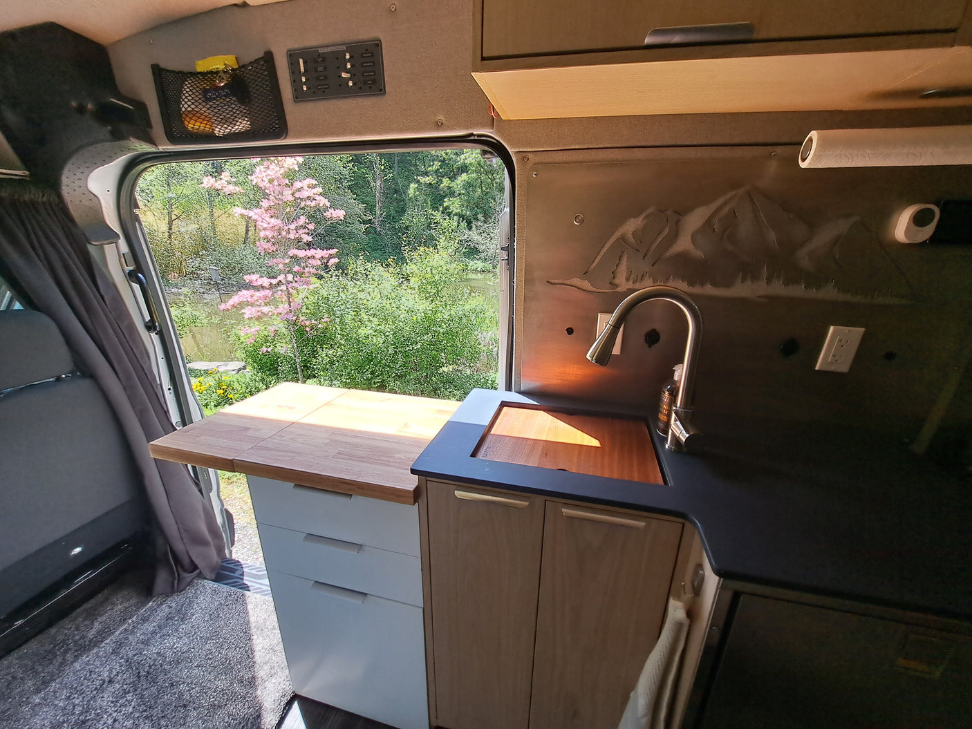 Experience nature with this RV rental! Camper van kitchen featuring a sink, counter, and storage, overlooking a scenic lake and trees.