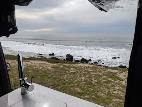 Oceanfront RV rental view: waves, rocky shore, and an interior sink, perfect for a coastal van life adventure.