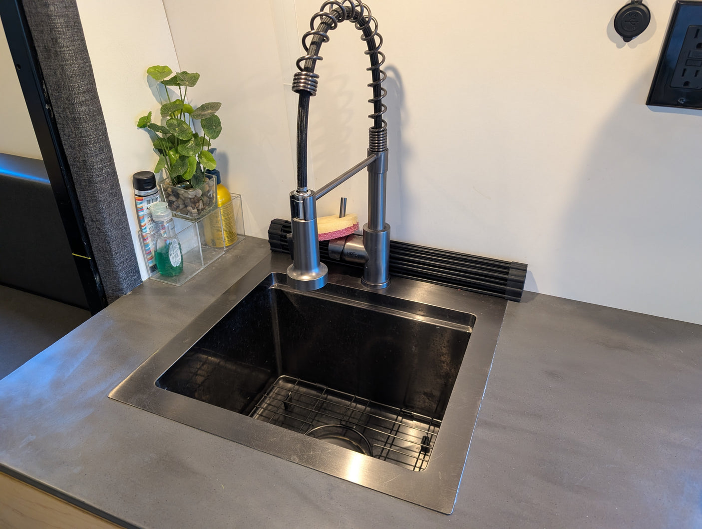 Modern rv rental kitchen area with a dark sink, industrial faucet, and grey counter. Features a drying rack, plant, and wall outlet.