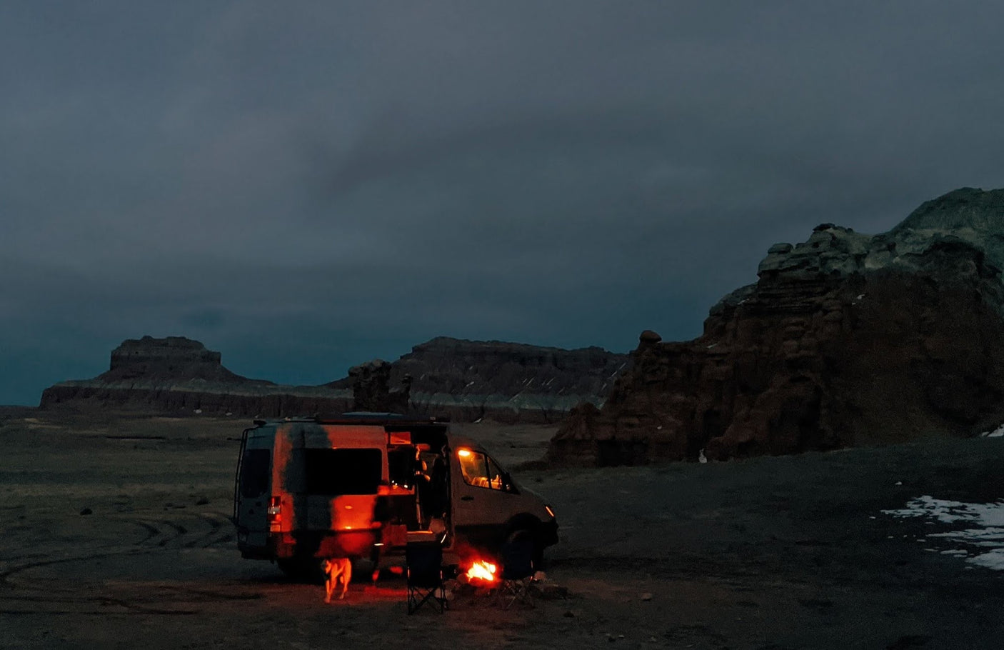 Dark night, an RV rental parked in a vast, rocky desert. A glowing campfire and dog add warmth to the scene.