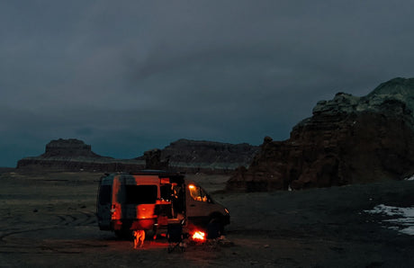 Dark night, an RV rental parked in a vast, rocky desert. A glowing campfire and dog add warmth to the scene.
