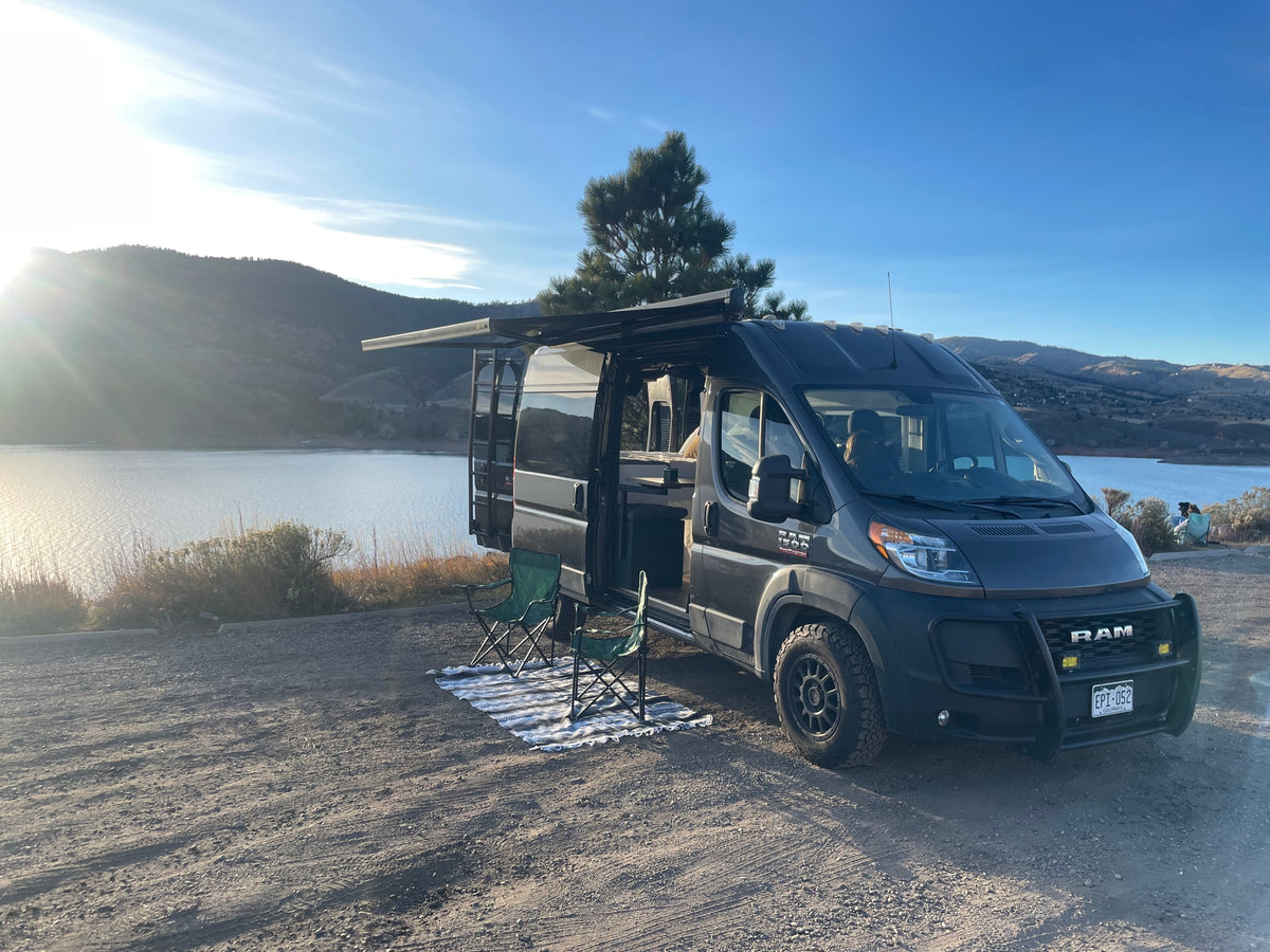 A dark gray camper van from an RV rental service parked lakeside with mountains in the distance, awning out and chairs set up. — photo 14