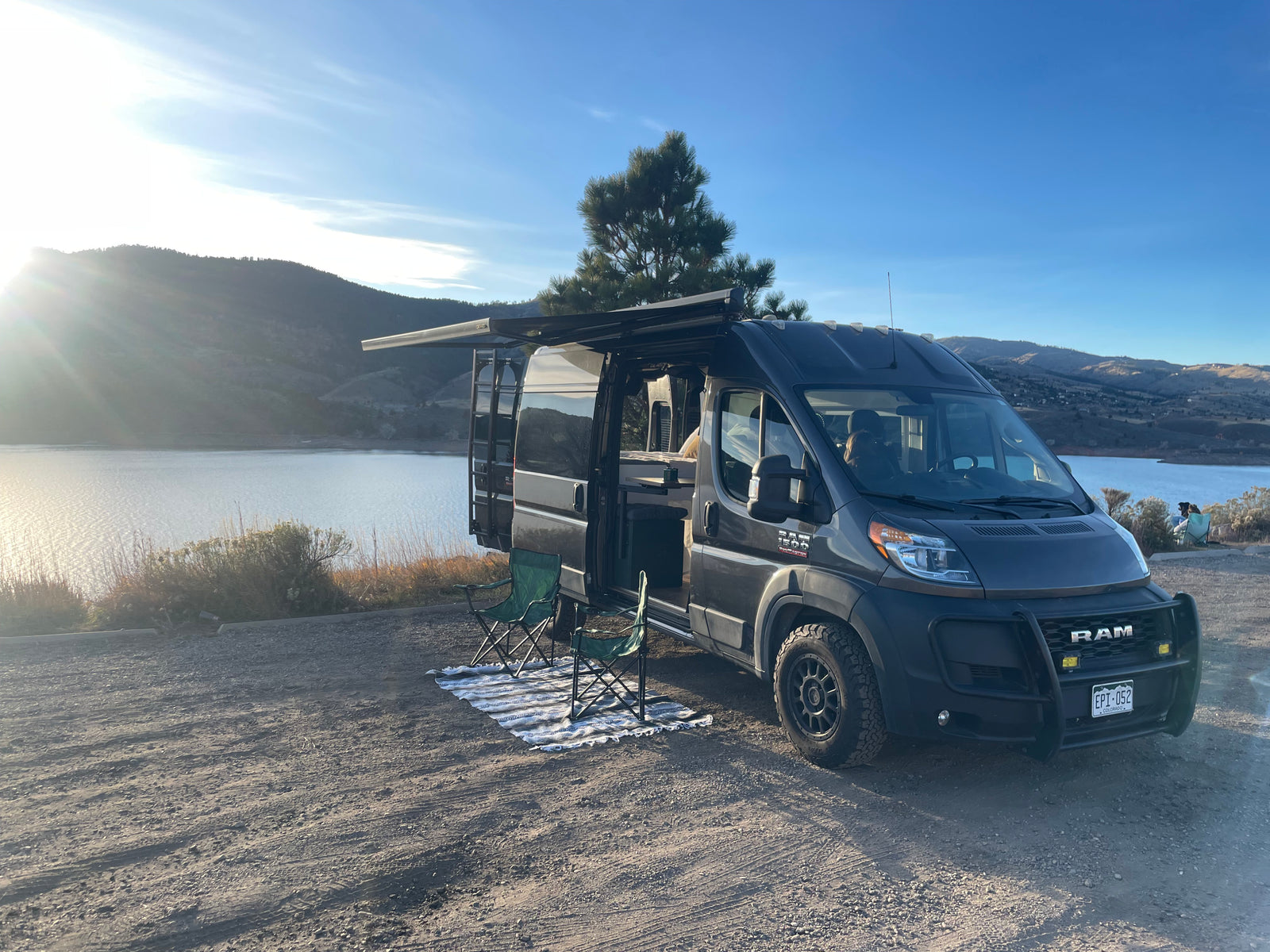 A dark gray camper van from an RV rental service parked lakeside with mountains in the distance, awning out and chairs set up.