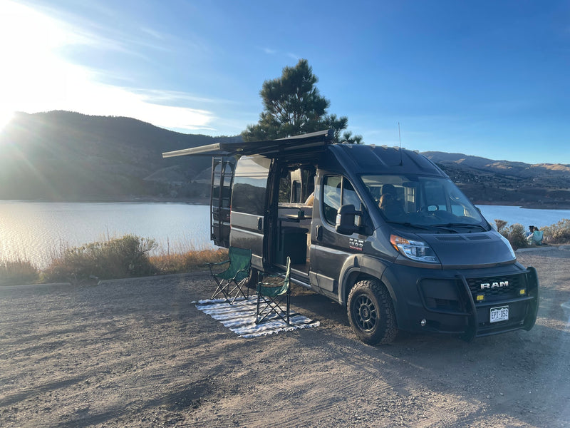A dark gray camper van from an RV rental service parked lakeside with mountains in the distance, awning out and chairs set up. — photo 14