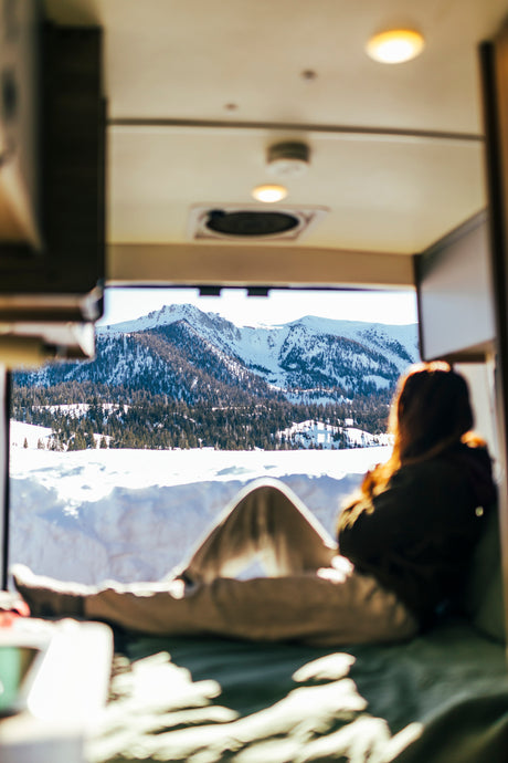 Enjoying a serene winter mountain view from inside an RV rental. A person relaxes on a bed, looking out at the snowy peaks and forests.