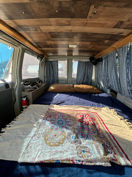 Cozy rv rental van interior with bed, wood ceiling, patterned curtains, fire extinguisher, and fishing rods for adventure.