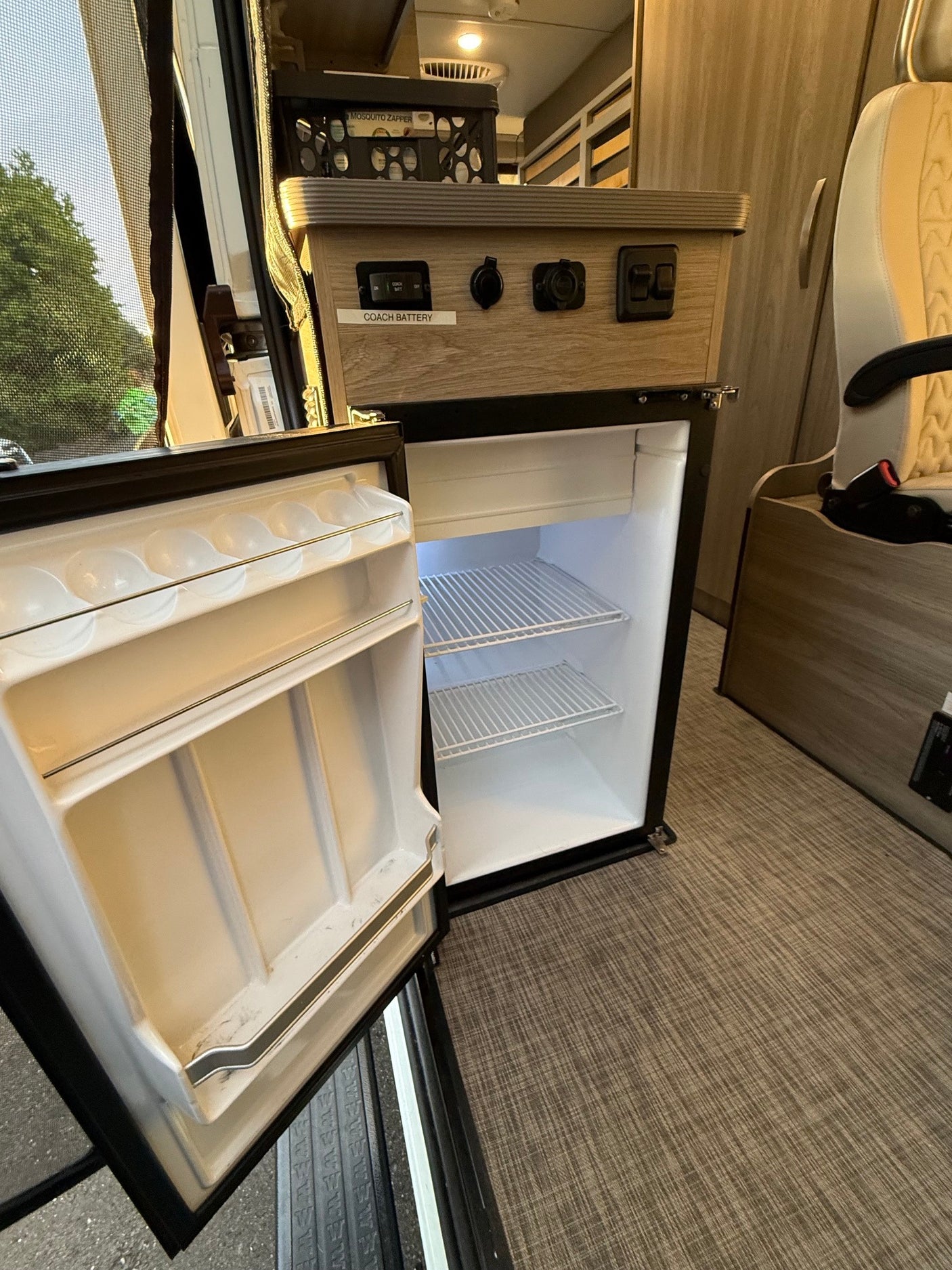 An open, empty mini-fridge in an RV rental, showing interior shelves, door storage, and a control panel with power outlets above.