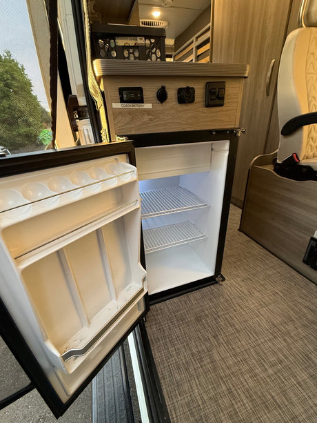 An open, empty mini-fridge in an RV rental, showing interior shelves, door storage, and a control panel with power outlets above.