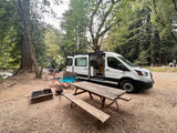 A white camper van, an RV rental, is perfectly set up at a serene forest campsite with a river, picnic table, and fire pit.
