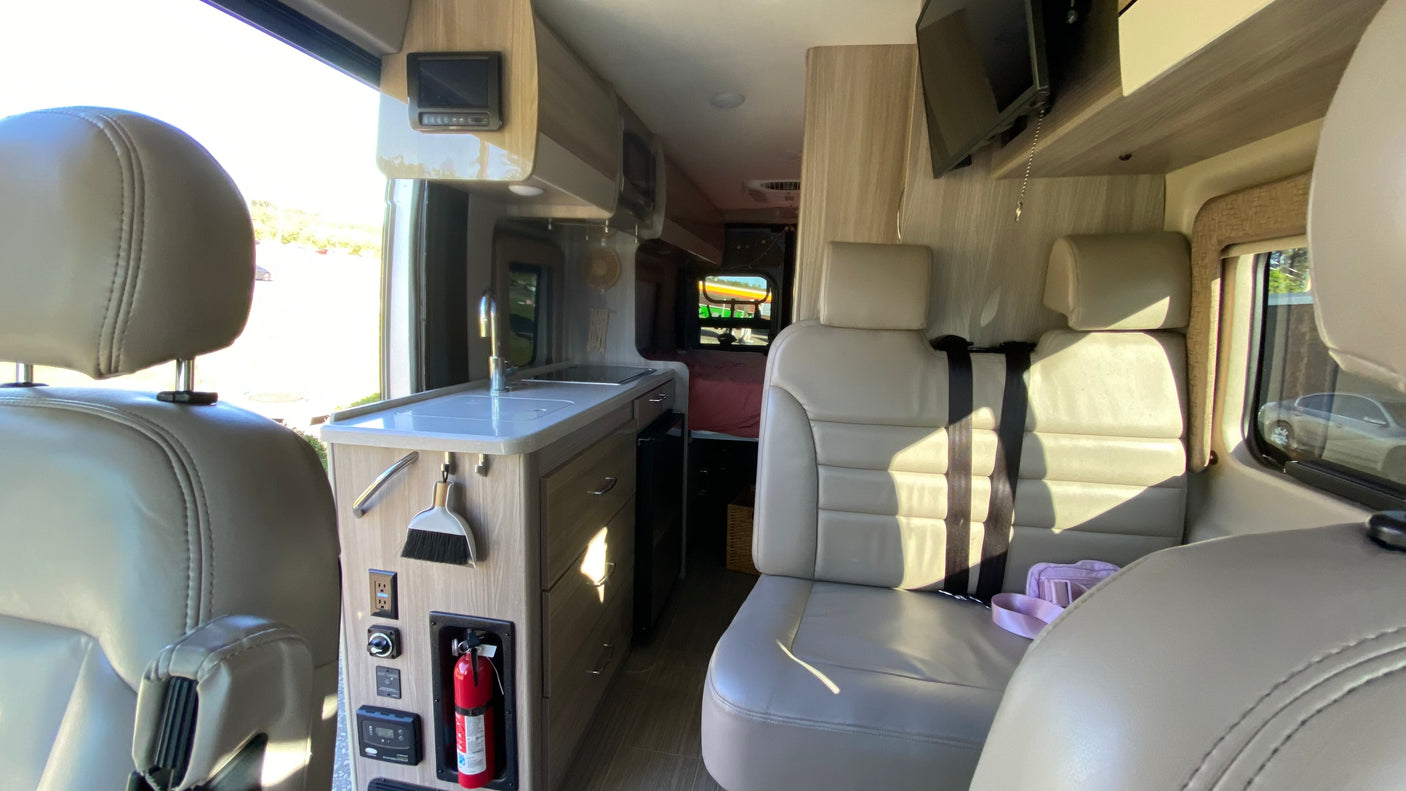 Inside a modern RV rental: light-colored seating, a functional kitchen with sink, overhead cabinets, and entertainment amenities.