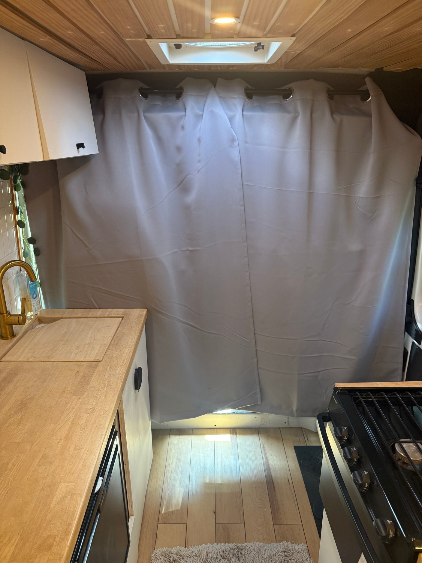 RV rental campervan kitchen with wooden ceiling, light counters, gold faucet, stove, and light gray privacy curtains.