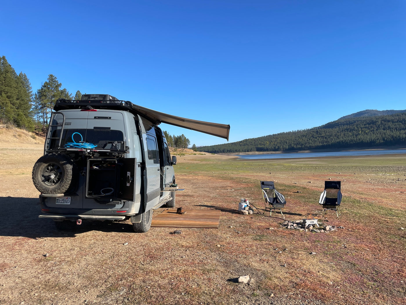 Grey RV rental camper van with awning, camping chairs, and a fire pit set up by a dried-up lake bed. — photo 5