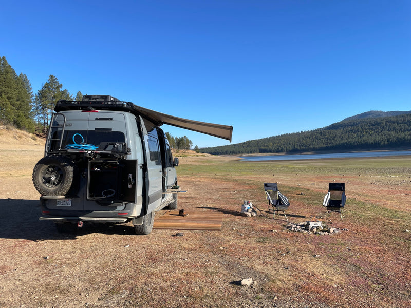 Grey RV rental camper van with awning, camping chairs, and a fire pit set up by a dried-up lake bed. — photo 5