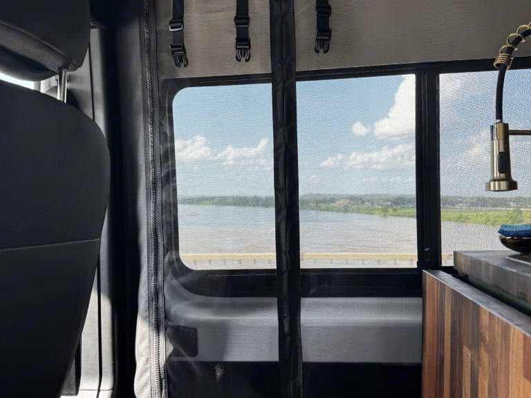 Inside an RV rental, enjoy expansive river views through a screened window, with a modern kitchen faucet and a comfortable seat.
