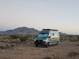 A grey RV rental van with a roof rack is parked in a vast desert landscape, with rugged mountains in the distance under a twilight sky.