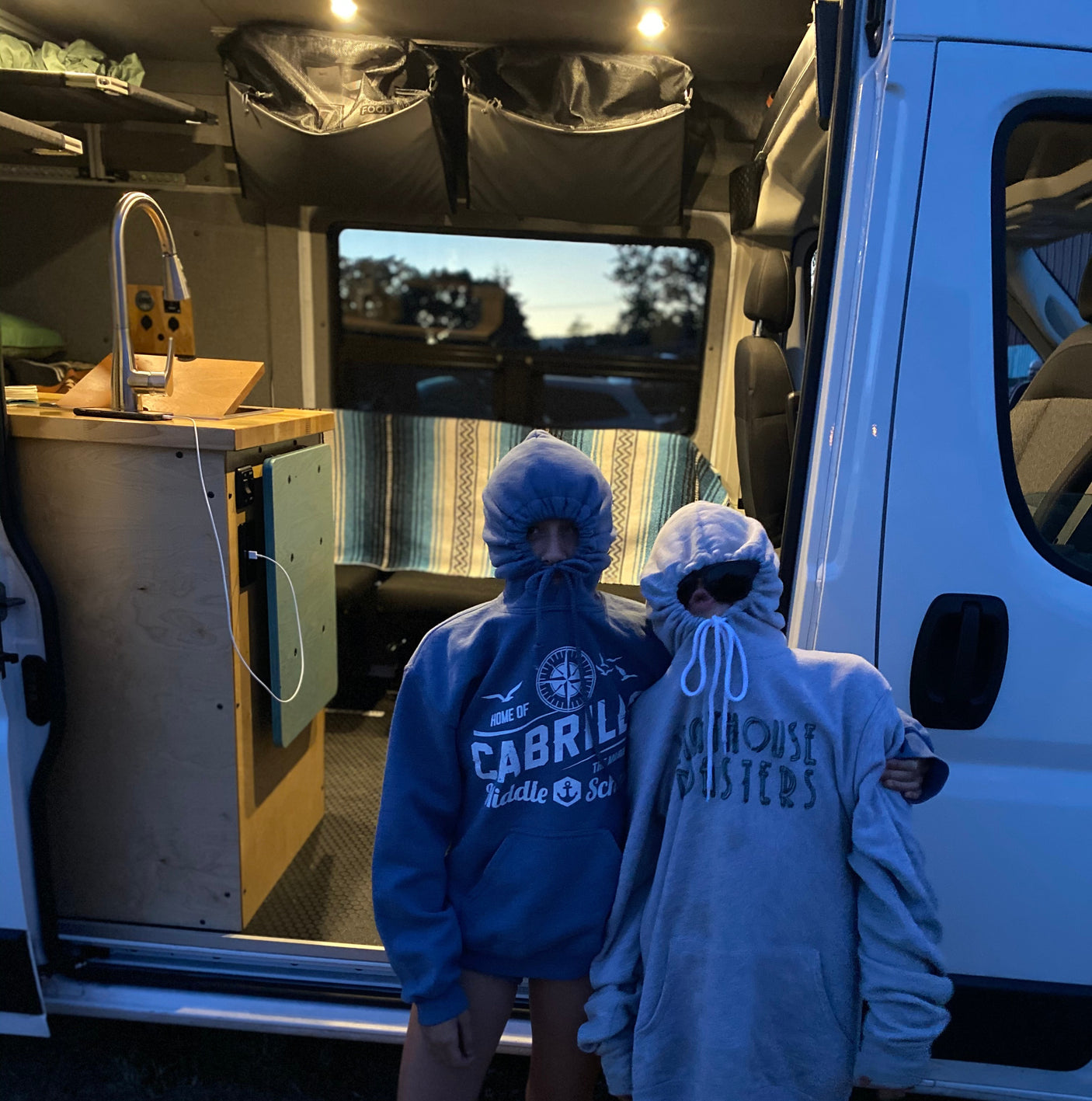Two people in hoodies stand in an RV rental van, showcasing its cozy interior with a sink, storage, and bench.