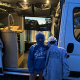Two people in hoodies stand in an RV rental van, showcasing its cozy interior with a sink, storage, and bench.