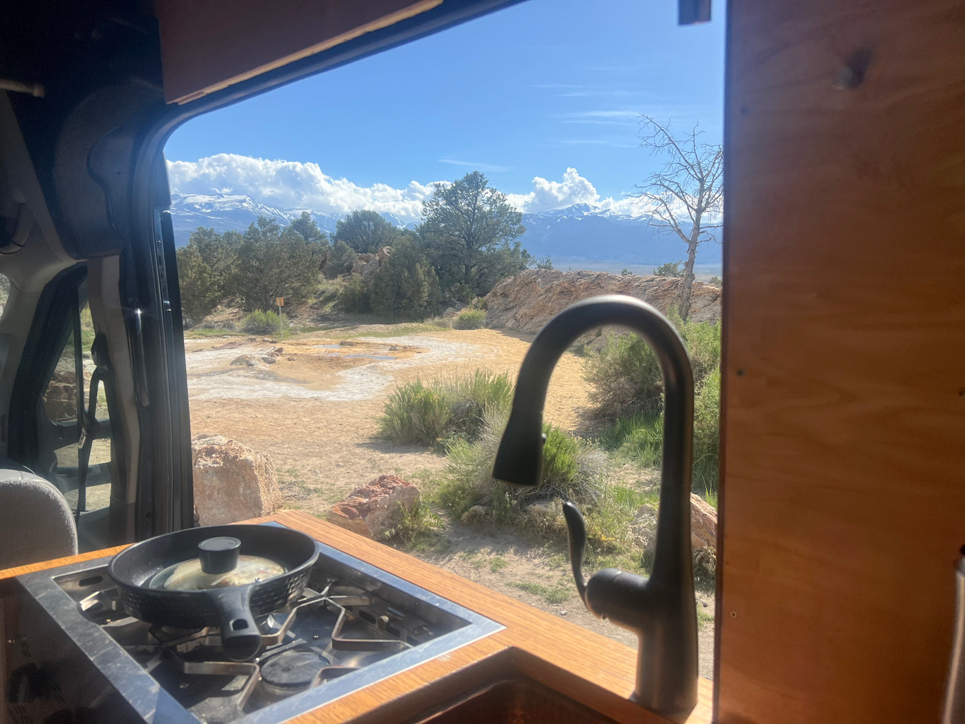 RV rental: panoramic views of hot springs & snowy mountains from inside a camper van's kitchen while cooking.