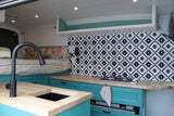 This RV rental interior showcases a cozy custom kitchenette with teal cabinets, wood counters, and a stylish backsplash, next to a raised bed.