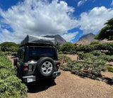 Dark grey Jeep with a Thule Tepui rooftop tent, an rv rental parked on a dirt path with lush trees and mountains.