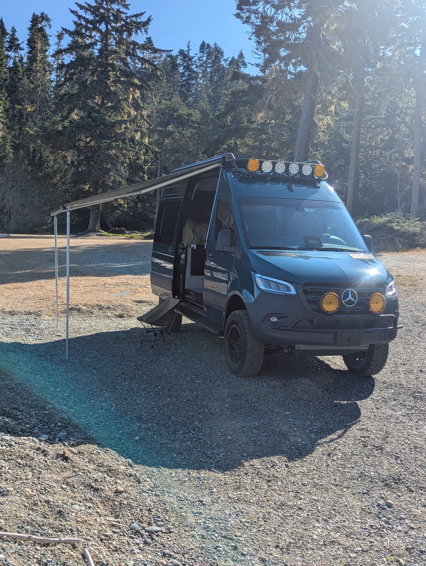 Adventure-ready Mercedes Sprinter RV rental parked on gravel with awning extended, surrounded by a sunny forest.