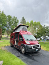 Red pop-top camper van (RV rental) parked on a wet driveway at a green campsite under cloudy skies.