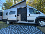 This silver RV rental is seen with its pop-top open, side door screen, and outdoor mat on the grass, ready for camping.