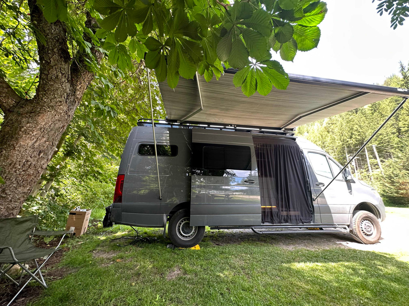 A gray **rv rental** van is parked at a grassy campsite with an open side door, extended awning, and folding chair.