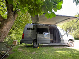 A gray **rv rental** van is parked at a grassy campsite with an open side door, extended awning, and folding chair.