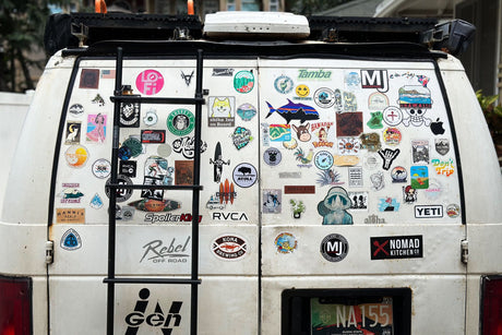 The back of a white RV rental van, adorned with a ladder and countless Hawaii-themed and outdoor adventure stickers.