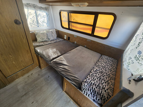 Cozy RV rental interior featuring a bed with patterned cushions, wooden walls, and windows with amber shades.