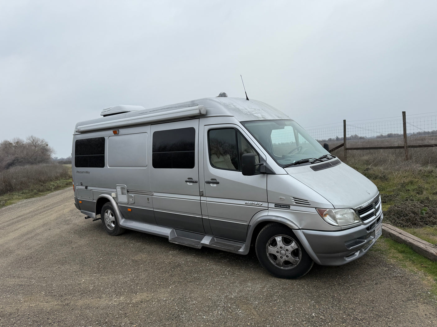 This silver Pleasure-Way Sprinter 2500 RV rental is ready for adventure, parked on a gravel road under an overcast sky.