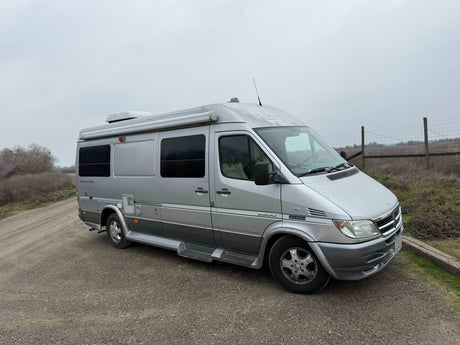 This silver Pleasure-Way Sprinter 2500 RV rental is ready for adventure, parked on a gravel road under an overcast sky.