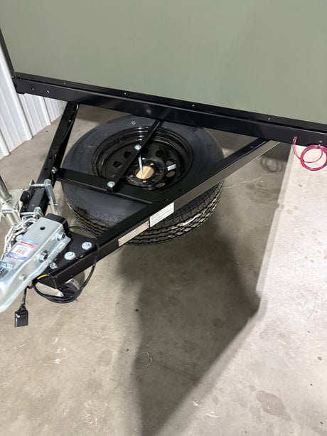Front section of an RV rental trailer, showcasing the hitch, black chassis, and spare tire mounted underneath on a concrete floor.