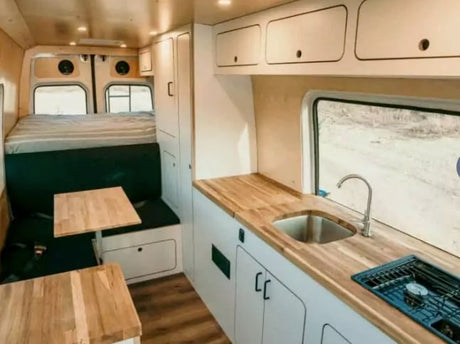 Interior of an RV rental showing a kitchen with sink and stove, dining bench, and bed, all with clean modern design.