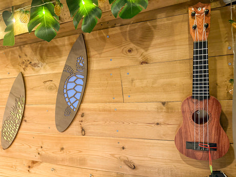 Wooden wall with a ukulele, two surfboard decorations (pineapple, turtle), and tropical string lights. Perfect RV rental vibe.